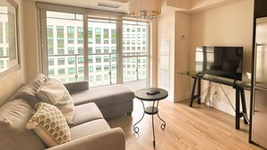 A modern living room with a sofa, a small round table, a TV on a glass stand, and large windows with a city view. - StayToronto - CN Tower & Front St Suites (Toronto)