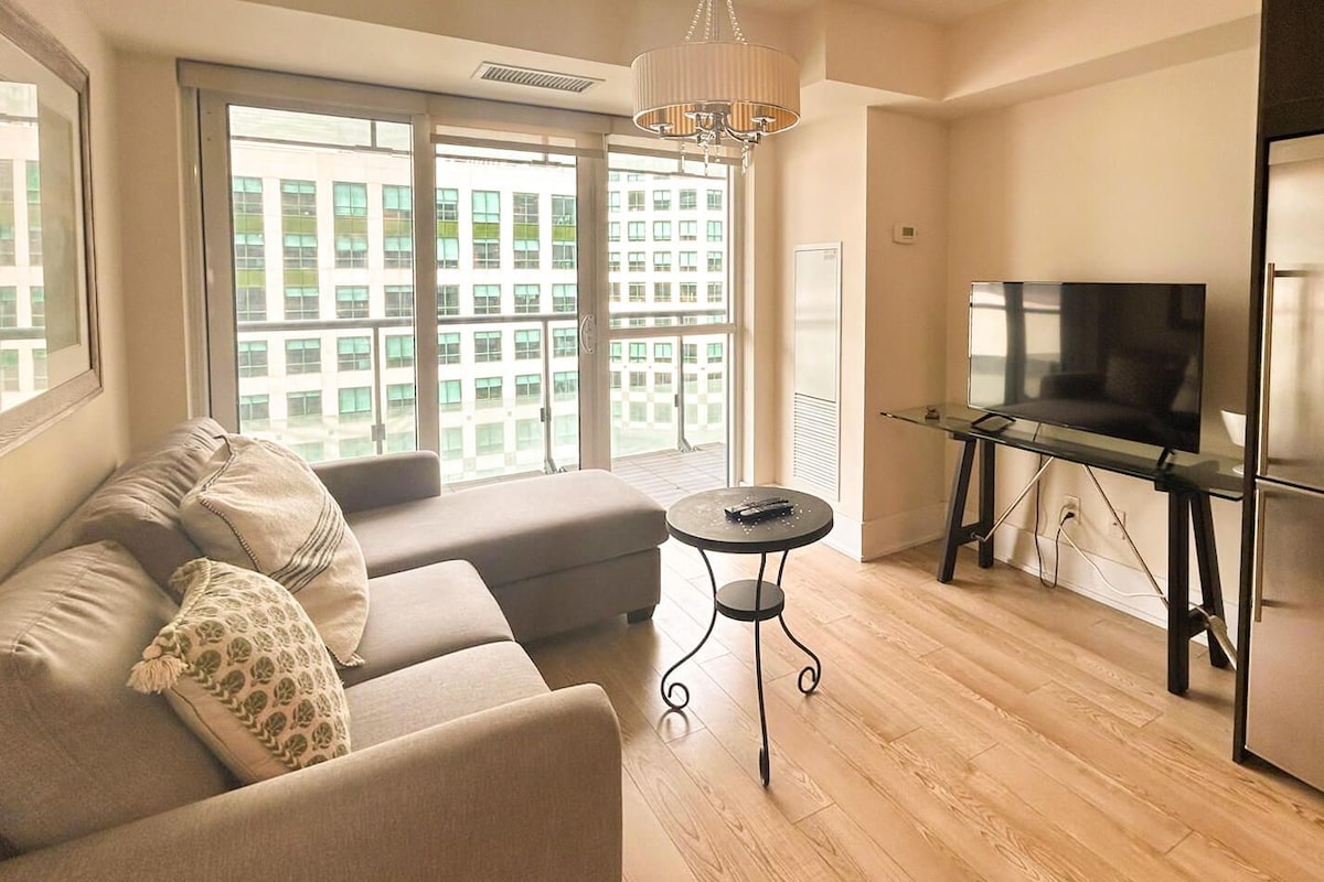 A modern living room with a sofa, a small round table, a TV on a glass stand, and large windows with a city view.
