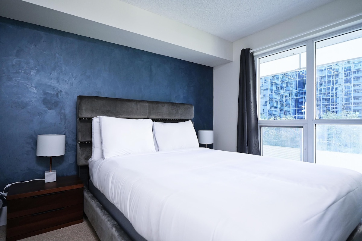 A bedroom with a large bed, a nightstand, a lamp, and a window with a view of buildings.