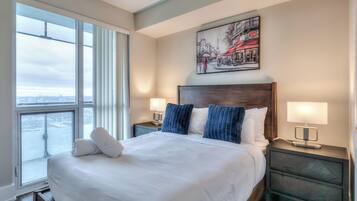 A hotel room with a large bed, two bedside tables with lamps, a cityscape view through a large window, and a framed picture on the wall.