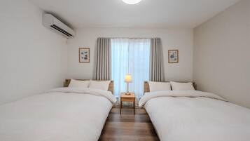 A hotel room with two beds, a bedside table with a lamp, an air conditioner, and a window with curtains.