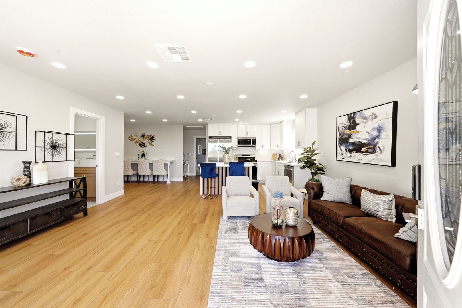 A modern living room with a wooden floor, a brown leather sofa, a coffee table, and a dining area with a white table and chairs.