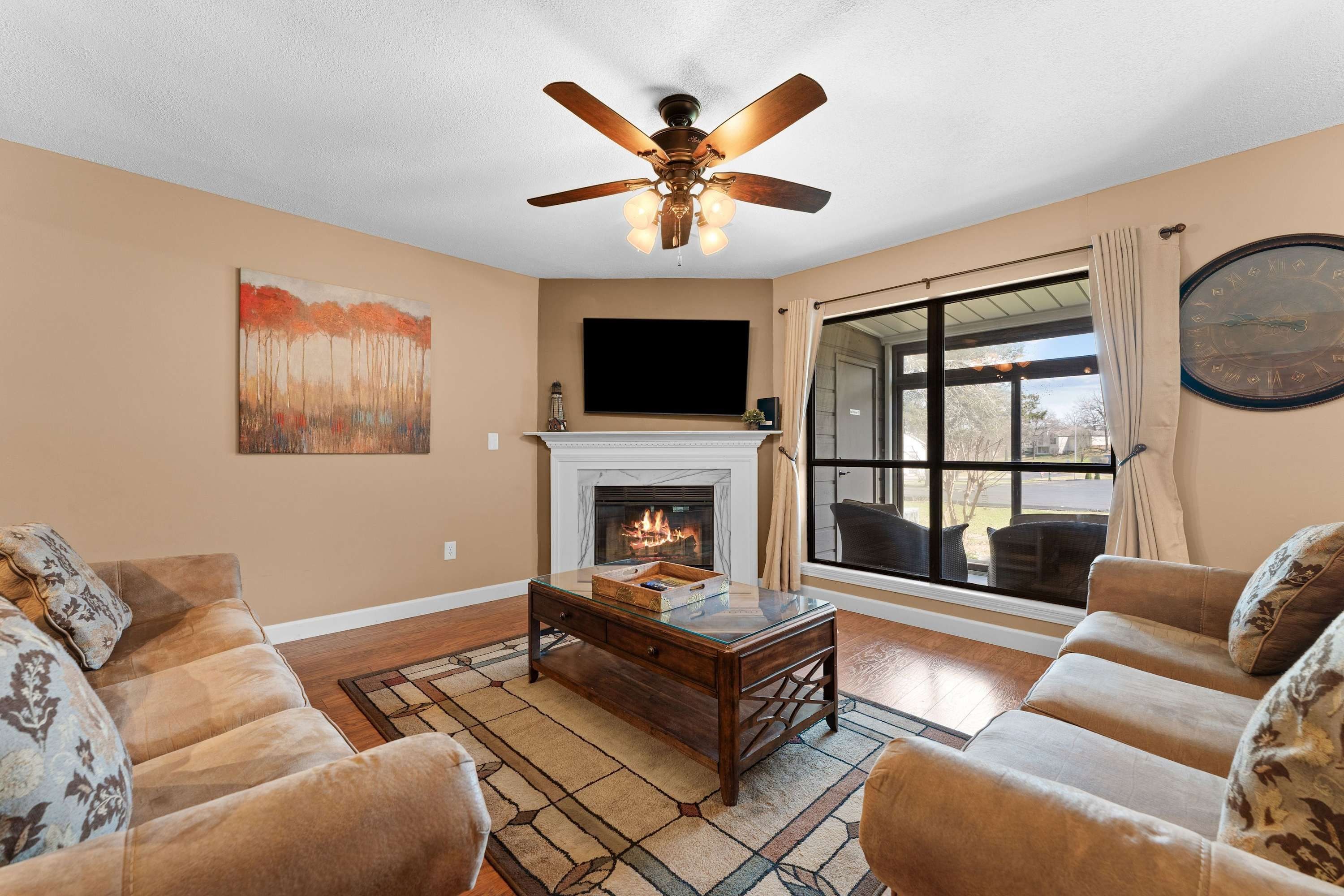 A living room with a fireplace, a ceiling fan, a large window, and a television mounted on the wall.