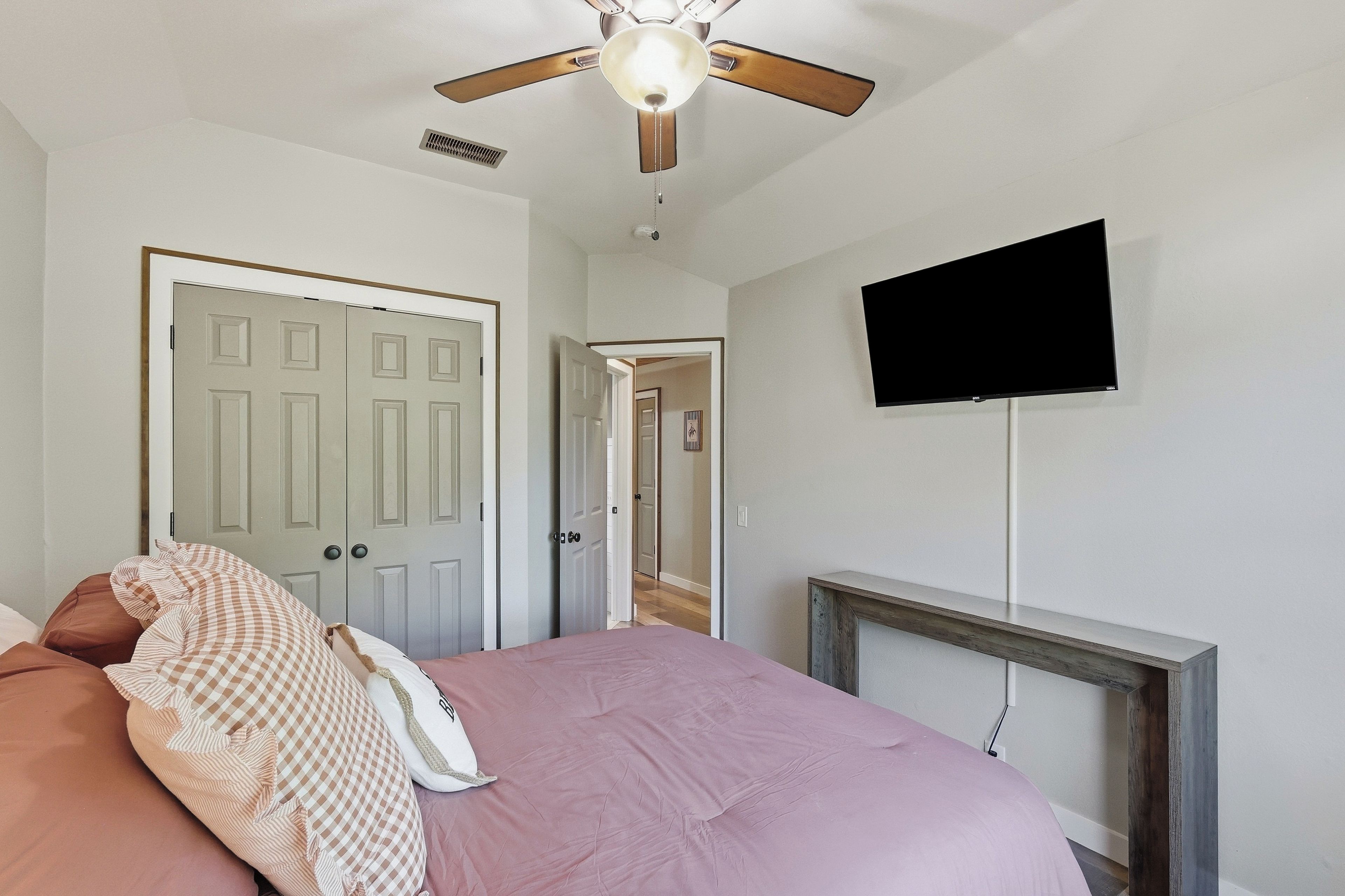 A bedroom with a bed, a ceiling fan, a television mounted on the wall, and a door.