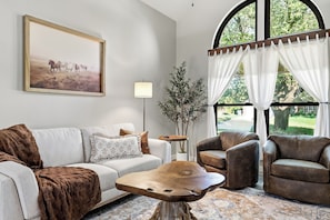 A living room with a sofa, armchairs, a coffee table, a painting, a lamp, and a large window with curtains. - Family Friendly Retreat, Fire Pit, Games, More (McKinney)