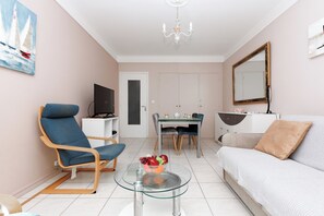A modern living room with a sofa, a blue armchair, a glass coffee table with fruit, a dining table with chairs, a TV on a stand, and a chandelier. - Upstays Cosy Calm 1 Bedroom with terrace and AC (Cannes)