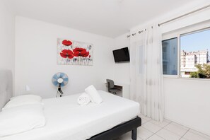 A hotel room with a bed, a desk, a chair, a TV, a window with a city view, and a painting on the wall. - Upstays Cosy Calm 1 Bedroom with terrace and AC (Cannes)