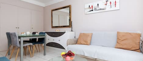 A modern living room with a sofa, a dining table with chairs, a glass coffee table with fruit, a mirror, and a painting on the wall.