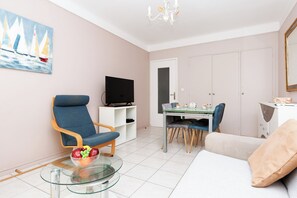 Ein Wohnzimmer mit blauem Sessel, einem Glastisch mit Früchten, einem Fernsehgerät auf einem Schrank, einem Esstisch mit Stühlen und einer Couch.
