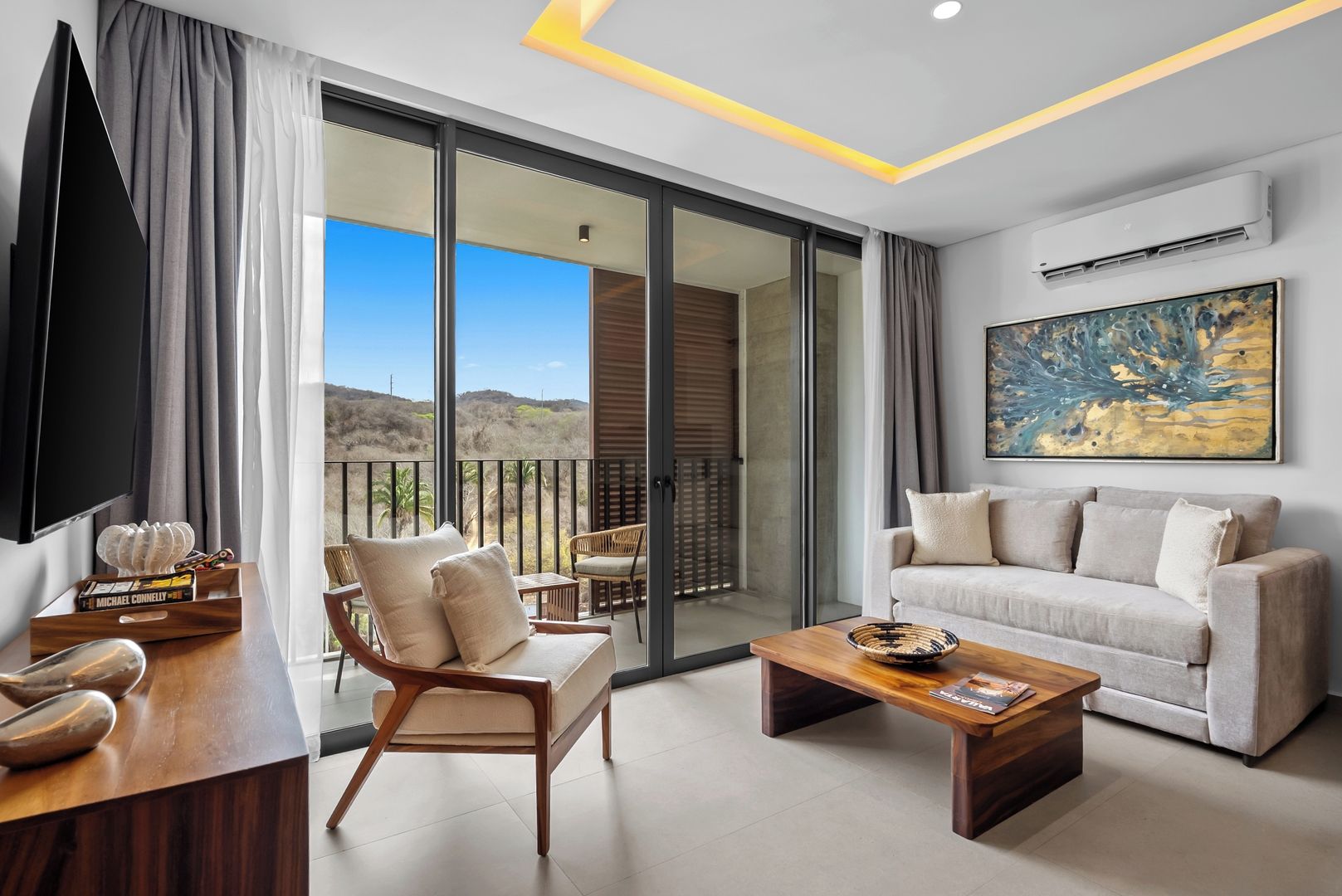 Una sala de estar moderna con un sofá, una mesa de centro y una televisión. Hay un balcón con vista al paisaje.