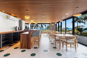 Ein modernes Restaurant mit Holztischen, hexagonalem Bodenmuster und großen Fenstern mit Blick auf den Ozean.
