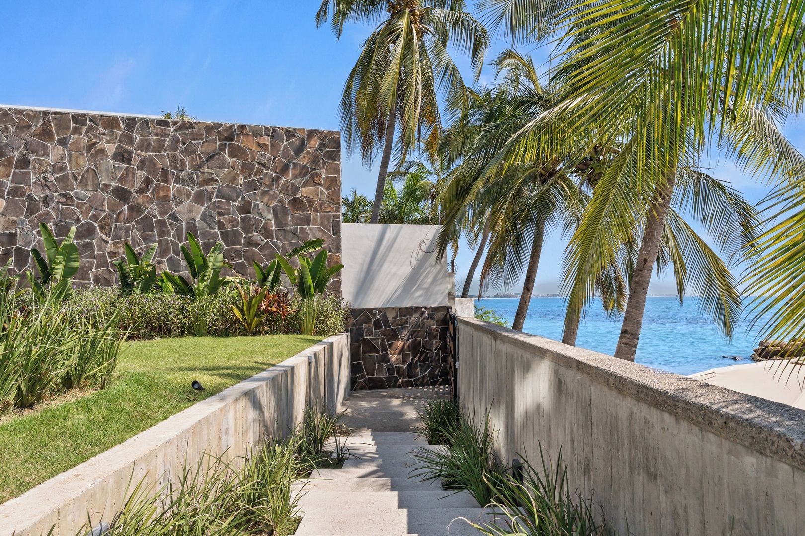 A pathway leading to a stone wall with a view of the ocean and palm trees.