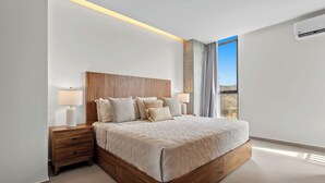 A modern bedroom with a wooden headboard, a large bed with a beige quilt, two bedside tables with lamps, and a view of the outdoors through a window. - Cozy Modern Condo with Scenic Balcony Views (Bucerías)