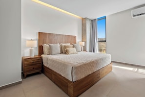 Ein modernes Schlafzimmer mit einem hölzernen Kopfteil, einem großen Bett mit einer beige farbenen Tagesdecke, zwei Nachttischen mit Lampen und Blick ins Freie durch ein Fenster.