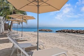 Ein Sitzbereich am Strand mit Sonnenschirmen und Liegestühlen, eine felsige Küstenlinie und ein freier Blick auf das Meer.
