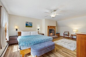A bedroom with a bed, a bench, a fireplace, a ceiling fan, and a painting on the wall.