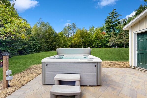 A hot tub with a step stool, surrounded by a patio with stone tiles, and a grassy lawn with trees in the background.