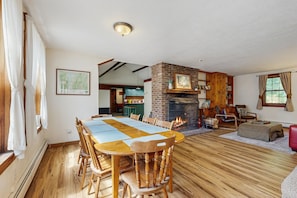Un área de comedor con una larga mesa de madera y sillas, una chimenea de ladrillo, y vistas a una cocina y comedor a través de un vano abierto.