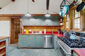 Una cocina con gabinetes de madera, un lavavajillas de acero inoxidable, una estufa a gas y ollas colgantes.