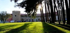 A building with a stone tower, a parking lot with cars, and a grassy area. - Palace Hotel & SPA Termas DE SAO Tiago (Penamacor)