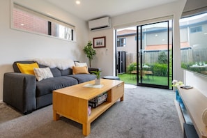 A modern living room with a grey sofa, a wooden coffee table, and a sliding glass door leading to a backyard with a patio. - Over The Moon | Quiet CBD w/ Courtyard (Christchurch)