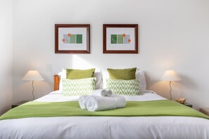 Une chambre d’hôtel avec un lit, des oreillers à motifs verts et blancs, un plaid vert, deux œuvres d’art encadrées accrochées au mur et des lampes de chevet.
