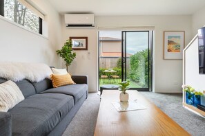 Un salon moderne avec un canapé gris, une table basse en bois et une vue sur un jardin, visible à travers une porte-fenêtre coulissante.