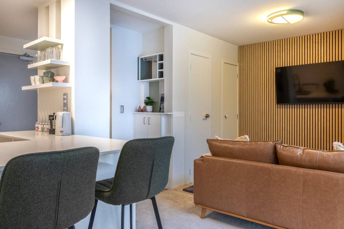 Ein modernes Wohnzimmer mit einem braunen Sofa und zwei grauen Sesseln, einem an der Wand montierten Flachbildfernseher sowie einer Küchenzeile mit weißen Schränken und einer weißen Arbeitsplatte.
