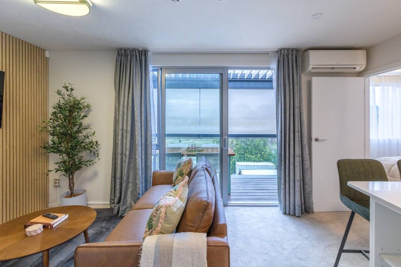 Un salon moderne avec un canapé, une petite table ronde, une plante en pot et une vue sur l’extérieur, visible à travers une porte vitrée.