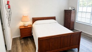 Une chambre à coucher avec un lit en bois, une table de chevet avec une lampe, un ventilateur de plafond, une fenêtre avec des stores et un radiateur.