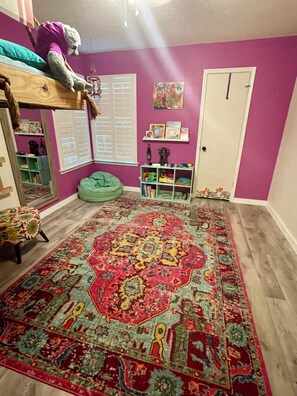 A room with a purple wall, a large patterned rug, a bunk bed, a bean bag chair, and a bookshelf.