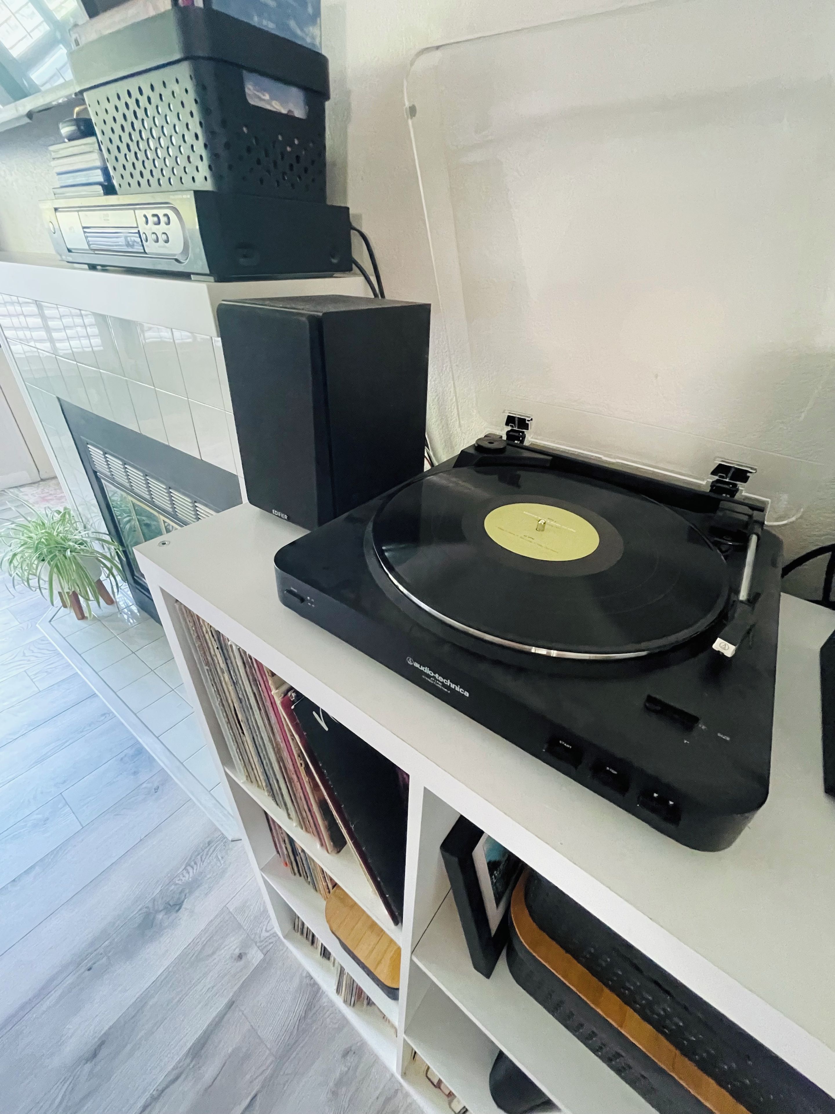 Une platine vinyle posée sur une étagère blanche, avec un haut-parleur et une plante en arrière-plan.