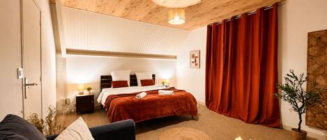 A hotel room with a wooden ceiling, a bed with red bedding, a round coffee table with candles, and a large window with red curtains.