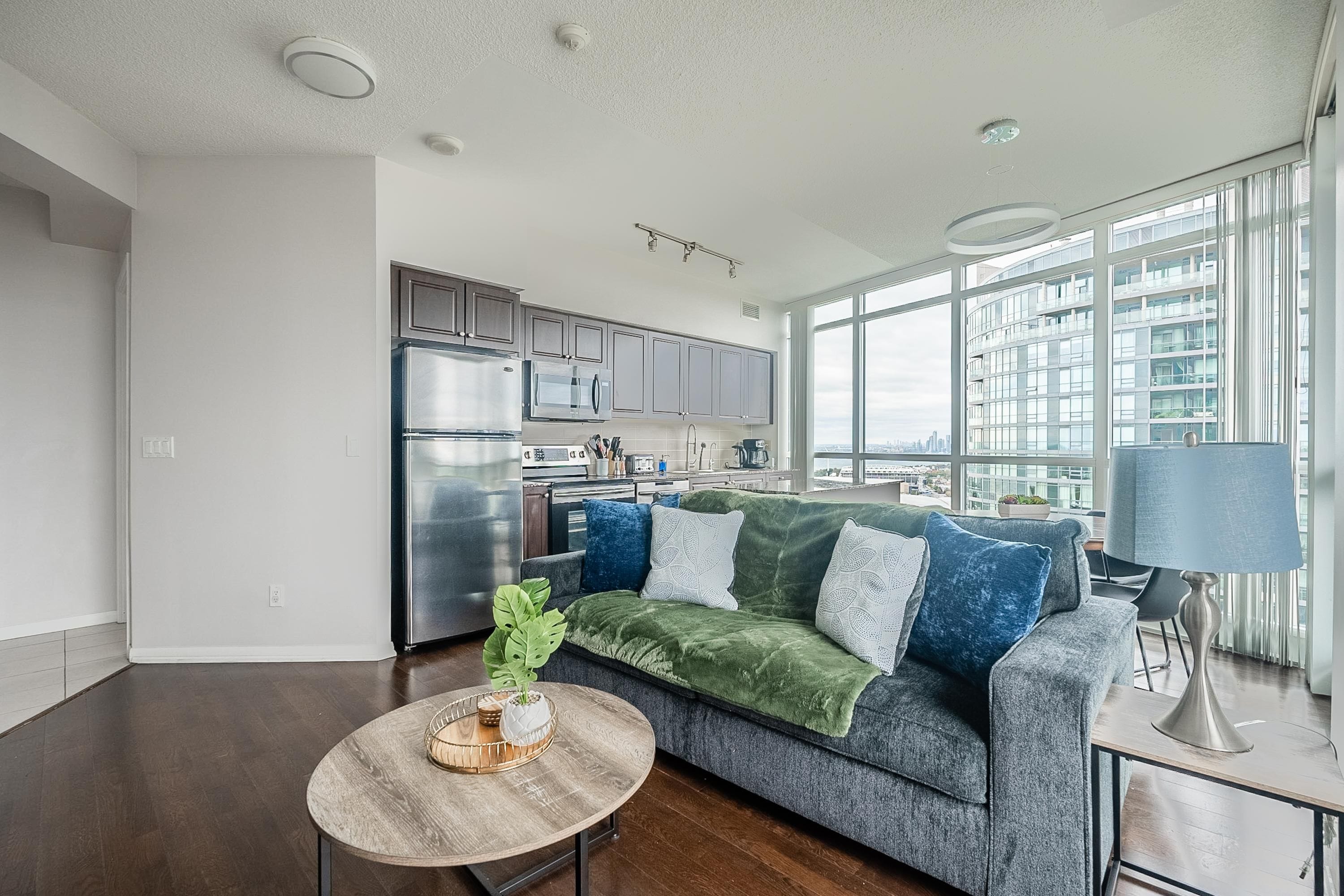 Een moderne woonkamer met een grijze bank, een houten salontafel en uitzicht op een stadsbeeld door grote ramen.