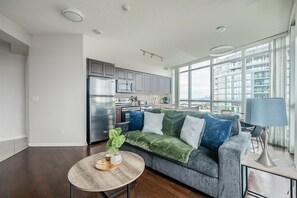Una sala de estar moderna con un sofá gris, una mesa de centro de madera y vistas a un paisaje urbano a través de amplias ventanas.