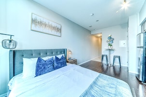 Quarto com cama, mesa de cabeceira, luminária na parede, quadro emoldurado e área de refeições com bancos.