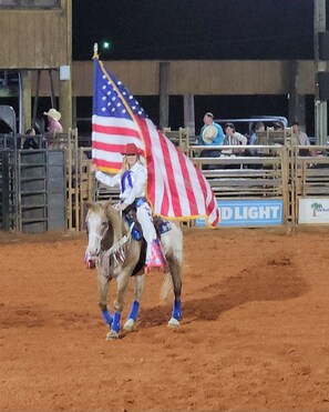 Une personne à cheval, portant un drapeau américain, dans une arène dont le sol est en terre et qui est entourée d’une clôture.
