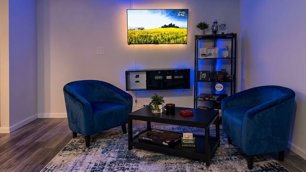 A modern living room with a flat-screen TV mounted on the wall, two blue armchairs, a coffee table, and a bookshelf with various items. -     Brand-New Luxury Home Spacious, Stylish & Your Home Away From Home (Tobyhanna)