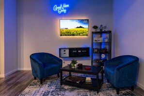 Ein modernes Wohnzimmer mit einem an der Wand befestigten Flachbildfernseher, zwei blauen Sesseln, einem Couchtisch und einem Bücherregal mit verschiedenen Gegenständen.