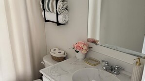 A bathroom with a white sink, a toilet, a shower curtain, a towel rack with towels, a basket with toilet paper, and a vase with flowers. -     Brand-New Luxury Home Spacious, Stylish & Your Home Away From Home (Tobyhanna)