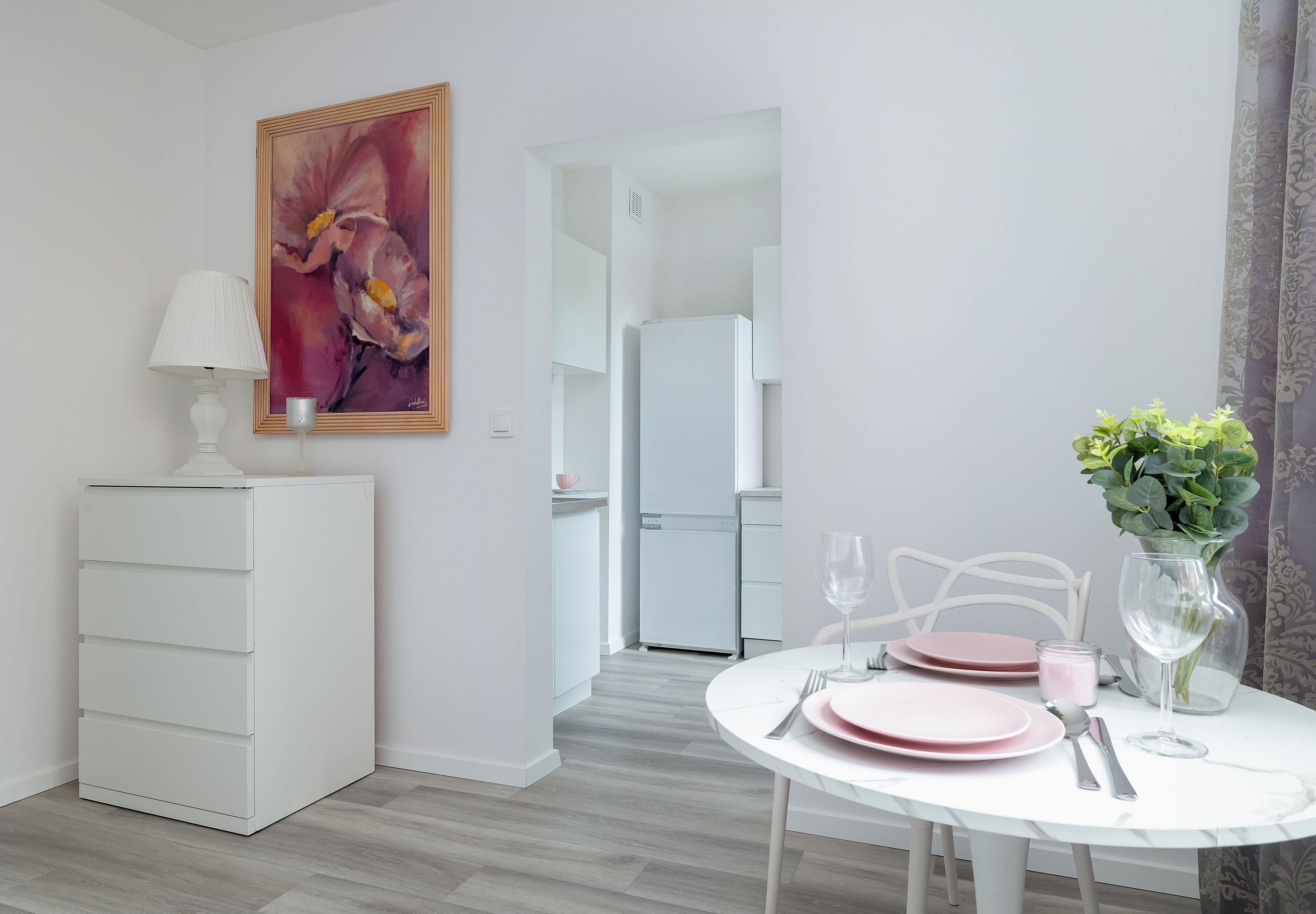 A modern dining area with a white table set for two, a white cabinet, and a painting on the wall.