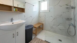 Une salle de bain avec un lavabo blanc, un miroir au-dessus, une douche équipée d’un pommeau de douche à main et une fenêtre laissant entrer la lumière naturelle.