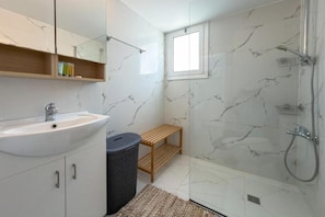 Ein Badezimmer mit einem weißen Waschbecken, einem Spiegel darüber, einer Dusche mit Duschbrause und einem Fenster für natürliches Licht.