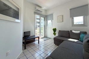 A modern living room with a sofa, a coffee table, and a television. There is a sliding glass door leading to an outdoor area. - Modern 2BR Beach Retreat at Harmony (Germasogeia)
