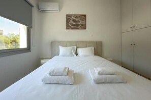A hotel room with a bed, two pillows, a window, an air conditioner, and a painting on the wall. - Modern 2BR Beach Retreat at Harmony (Germasogeia)