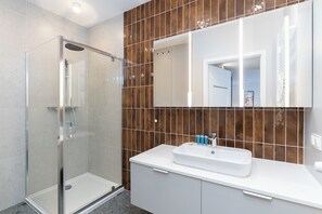 Ett modernt badrum med en duschavdelning med glasväggar, en vit tvättställ och en spegel med en väggbeklädnad av bruna kakelplattor bakom.