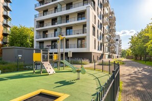 Un edificio residencial de varios pisos con balcones, un parque infantil con tobogán y estructura de escalada, y una calle pavimentada con una cerca.