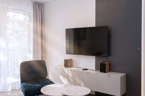 Ein modernes Wohnzimmer mit einem an der Wand befestigten Flachbildfernseher, einer weißen Kommode mit zwei Lautsprechern, einem blauen Sessel und einem runden weißen Couchtisch.