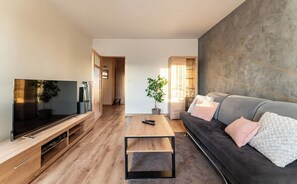 Ein modernes Wohnzimmer mit einem Flachbildfernseher, einem Holztisch, einem dunklen Sofa mit Kissen und einer großen Pflanze.