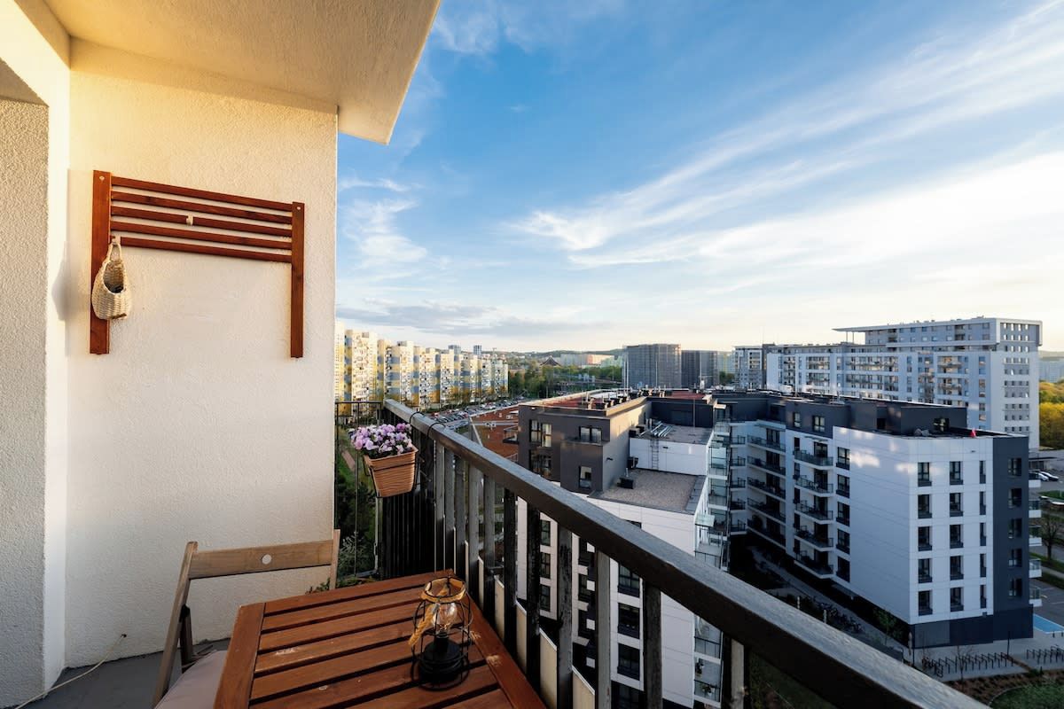 A balcony with a wooden table, a hanging basket with flowers, and a view of modern buildings.